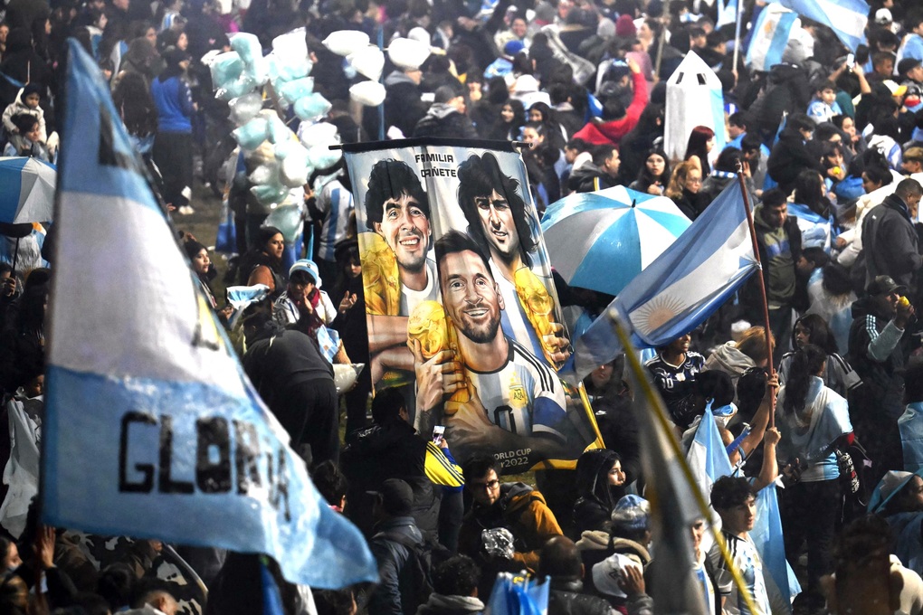 Người hâm mộ giương cao tấm băng rôn khắc họa ba huyền thoại bóng đá Argentina gồm Diego Maradona, Mario Kempes và Lionel Messi nâng cao cúp vàng World Cup, khi tập trung bên ngoài trung tâm huấn luyện của AFA tại Buenos Aires ngày 20/7. Ảnh: AFP